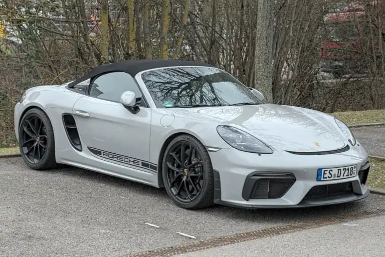 Chalk gray Porsche 718 Spyder with black soft top parked beside a tree line