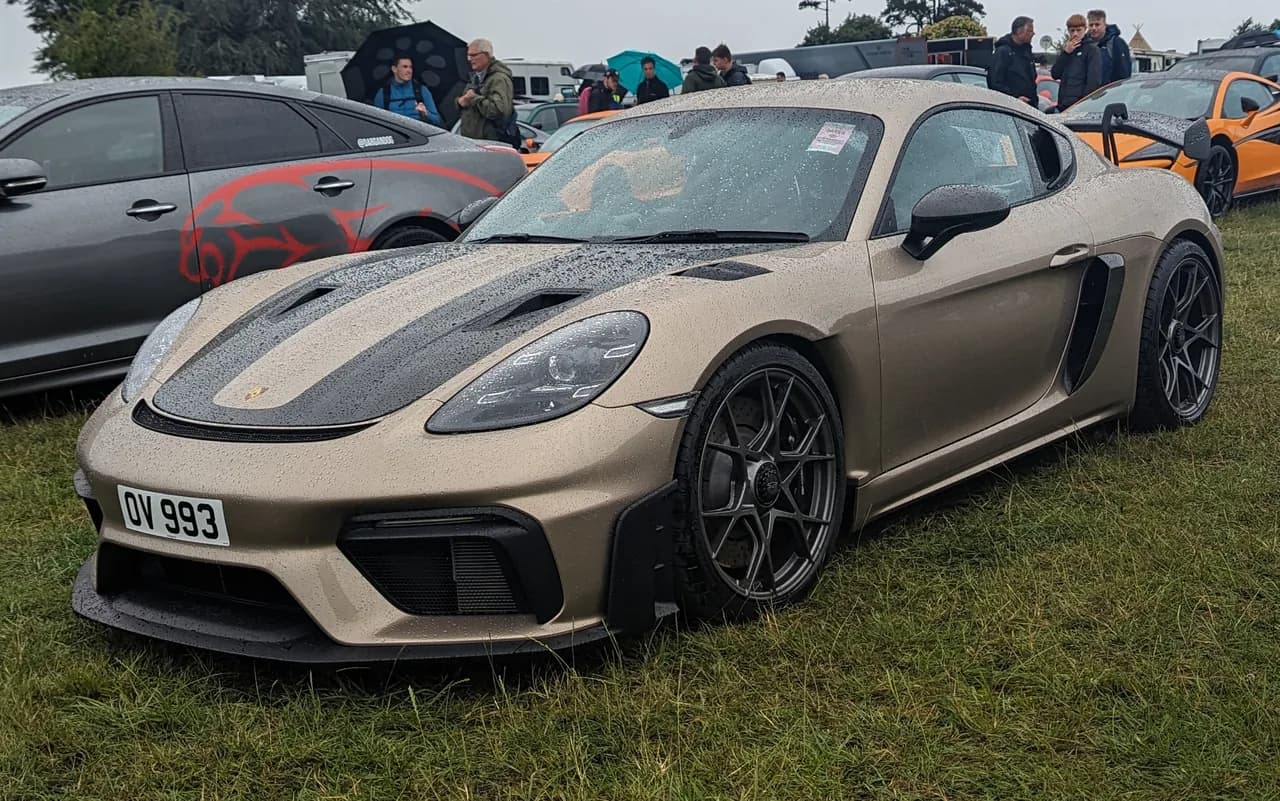 Wet beige Porsche 718 Cayman GT4 RS with a dark carbon-fiber hood, viewed from the front-left.