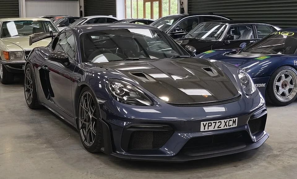 Dark blue Porsche 718 Cayman GT4 RS viewed from the front-left indoors, with dark hood panels and the fixed rear wing visible.