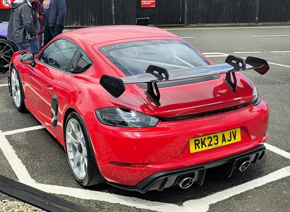Red Porsche 718 Cayman GT4 RS viewed from the rear-left, showing the fixed wing, rear diffuser, and twin exhaust outlets.