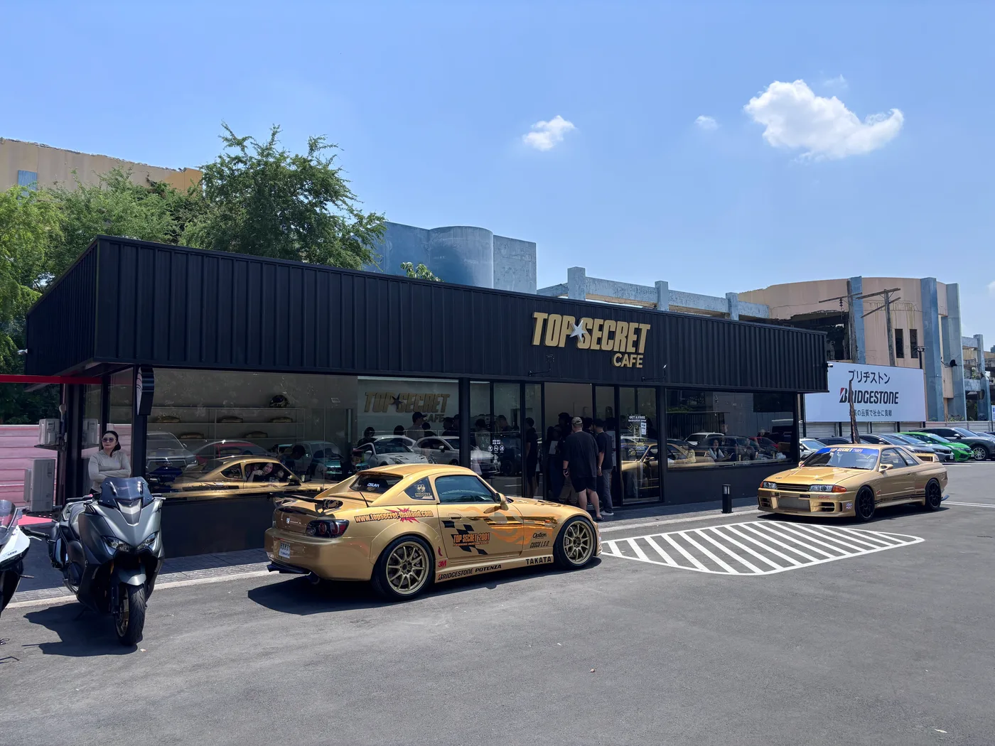 Two gold liveried Top Secret race cars parked outside the cafe entrance